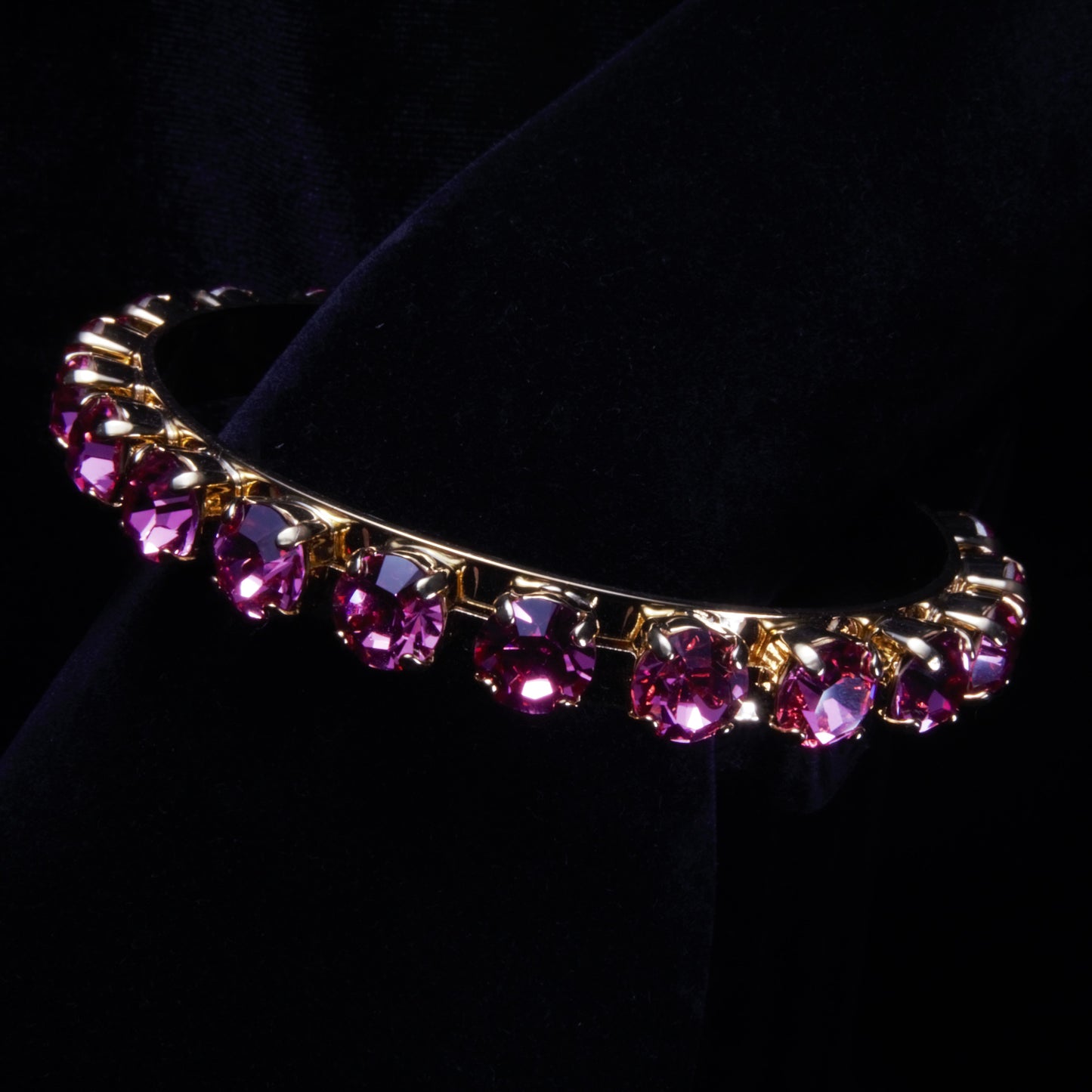 Set against a black background is a gold plated bangle bracelet with purple stone settings, it rests on the wrist of a model hand.